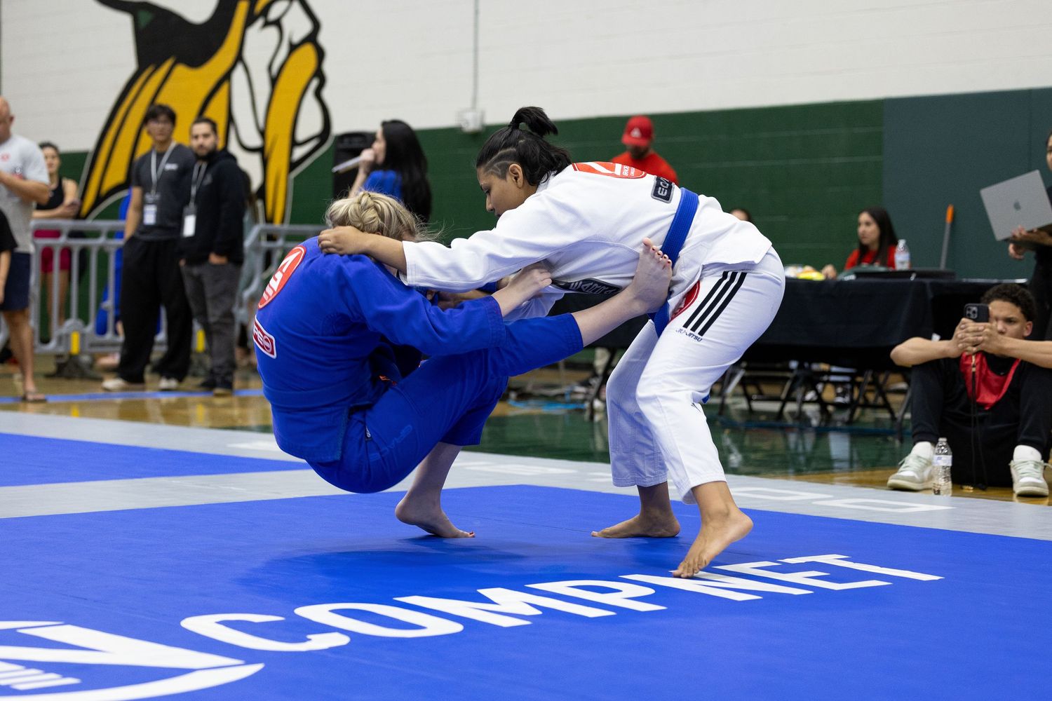 BJJ competitors grappling on blue competition mats during a tournament match.