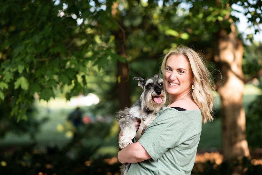 A person in a sage green shirt holds a small grey and white dog while standing in a sunlit wooded area at loose park kansas city.