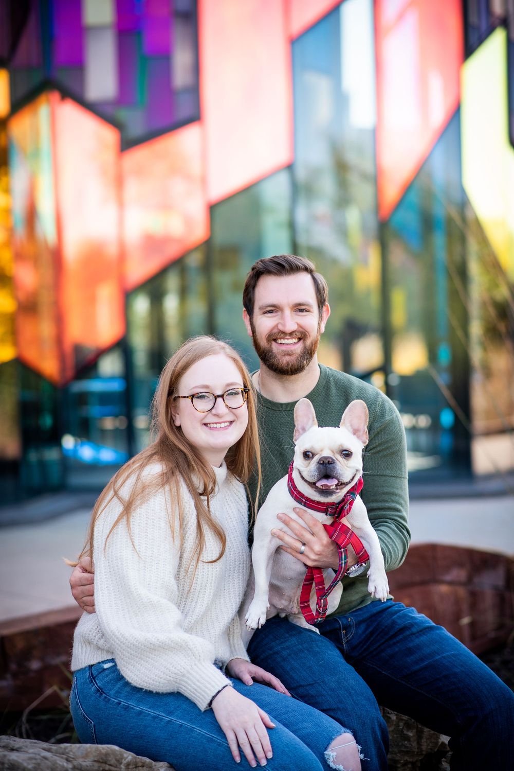 A couple sits together outdoors holding their French Bulldog in front of a colorful geometric wall display at prarie fire overland park ks.