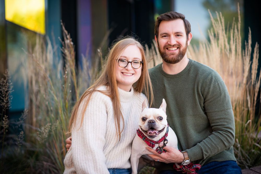 A smiling couple holds their happy white bulldog while posing outdoors among tall ornamental grasses at sunset.