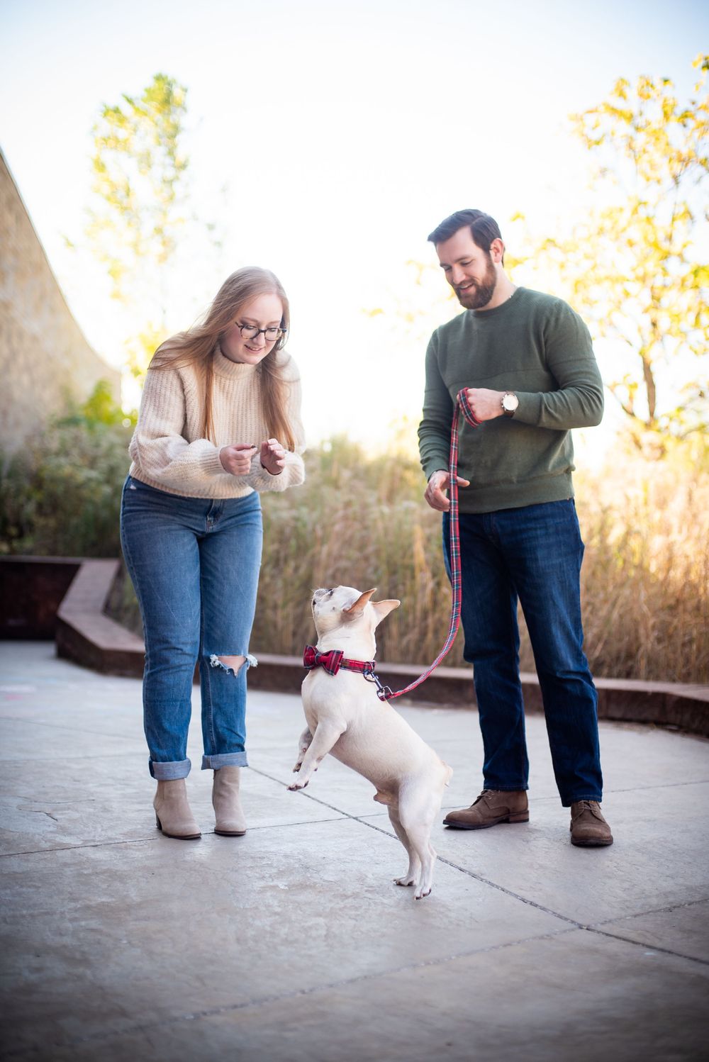 Two people in casual attire stand outdoors while training a small white dog on a leash during golden hour.