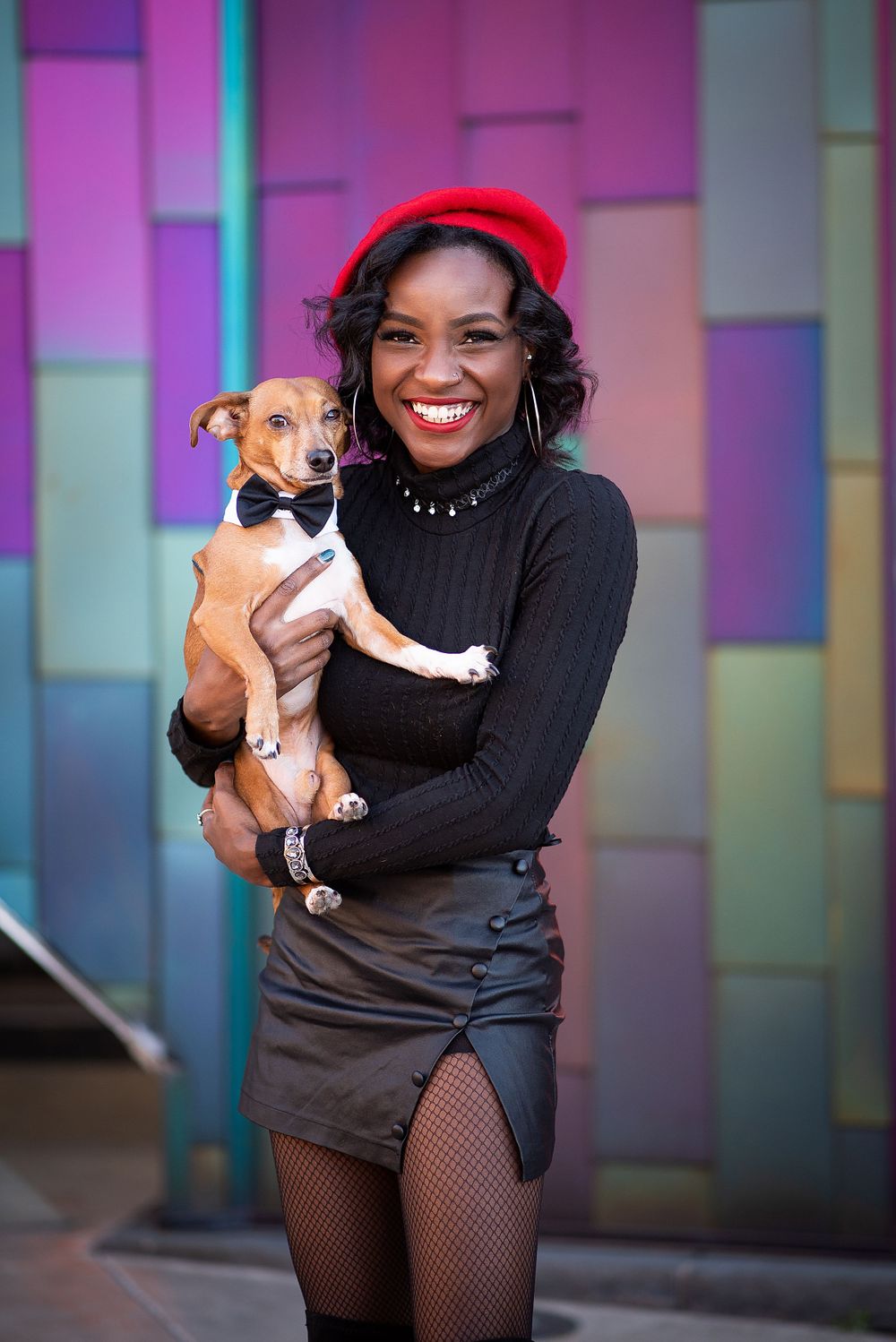 A person in a red beret and black dress holds a small brown and white dog wearing a bowtie against a colorful wall at prairie fire