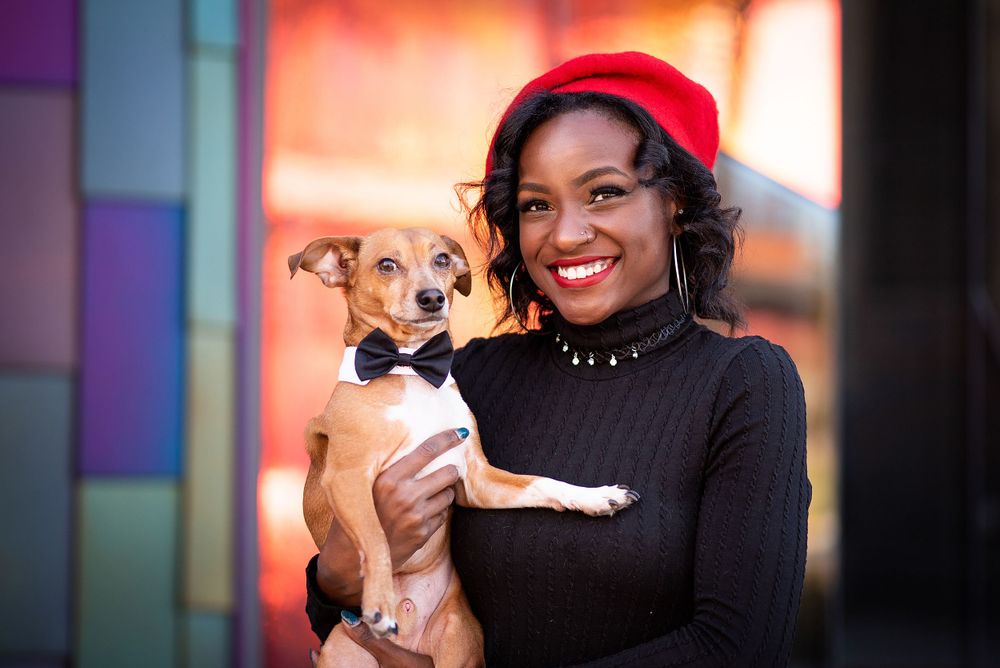 A small tan and white chihuahua mix dog is held lovingly while wearing a black bow tie collar against a colorful background in kansas city.