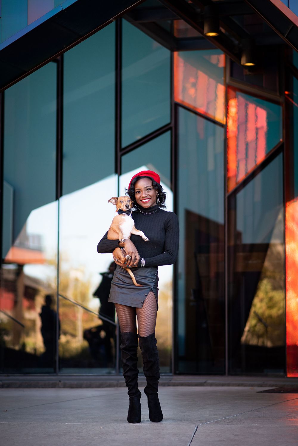 A person in a black sweater, red beret and knee-high boots holds a small chihuahua dog outside a modern glass building in overland park ks.