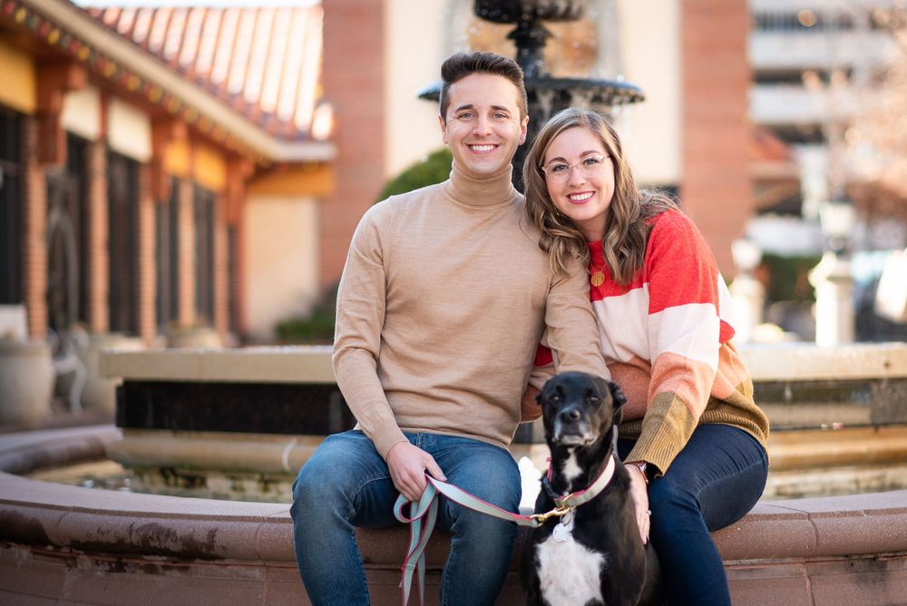A couple poses outdoors with their black and white dog on stone steps near a building with terracotta colored walls at the plaza district.