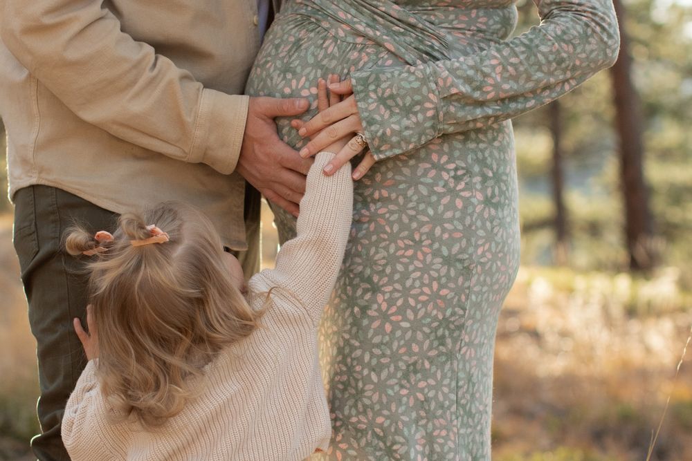 Small child in cream sweater reaches up to adult couple in beige and patterned clothing in outdoor autumn setting.