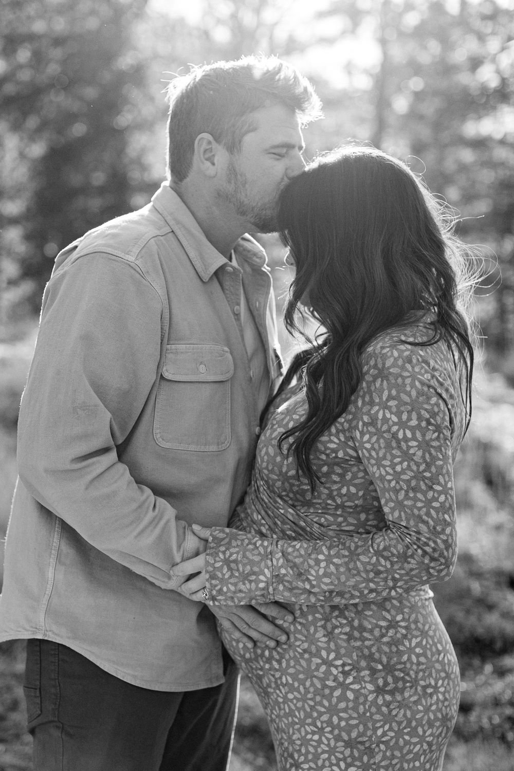 A tender black and white portrait of an expectant couple sharing an intimate moment outdoors in soft lighting.