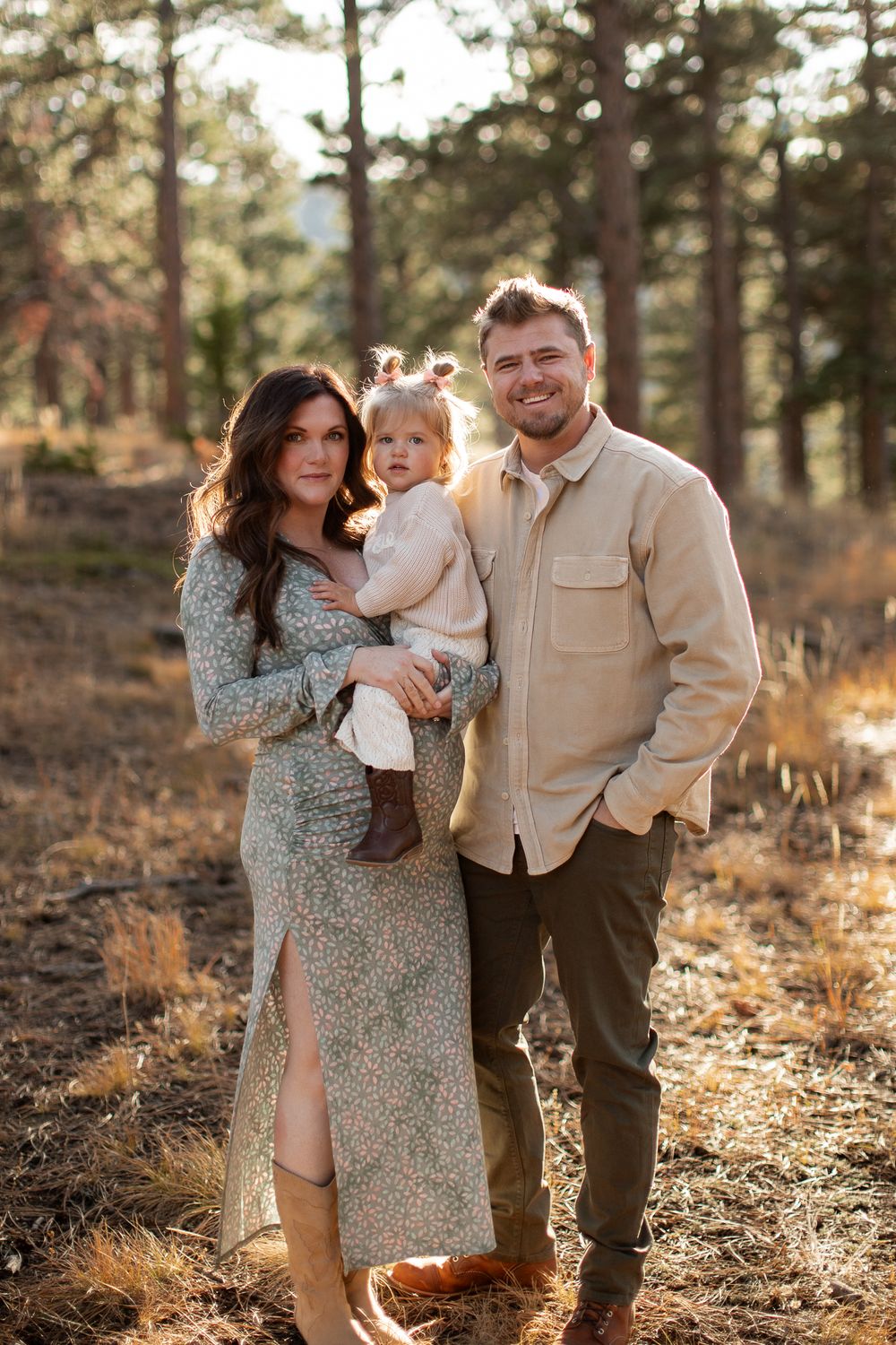 A family poses together in a sunlit forest setting wearing coordinated earth-toned outfits during golden hour.