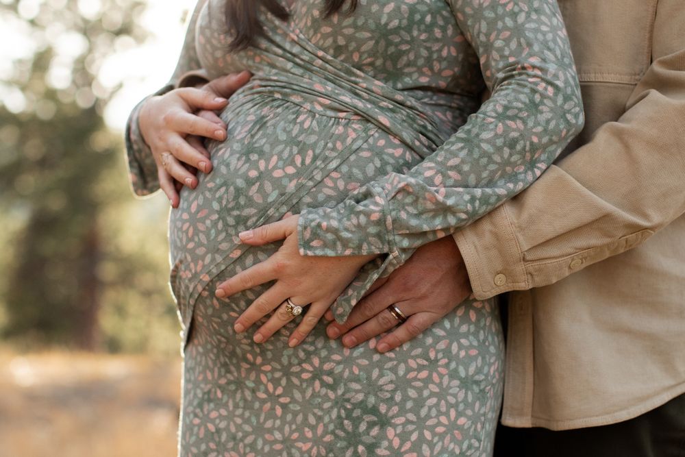 Close up shot of hands holding a pregnant belly wearing a green floral maternity dress with beige arms embracing.