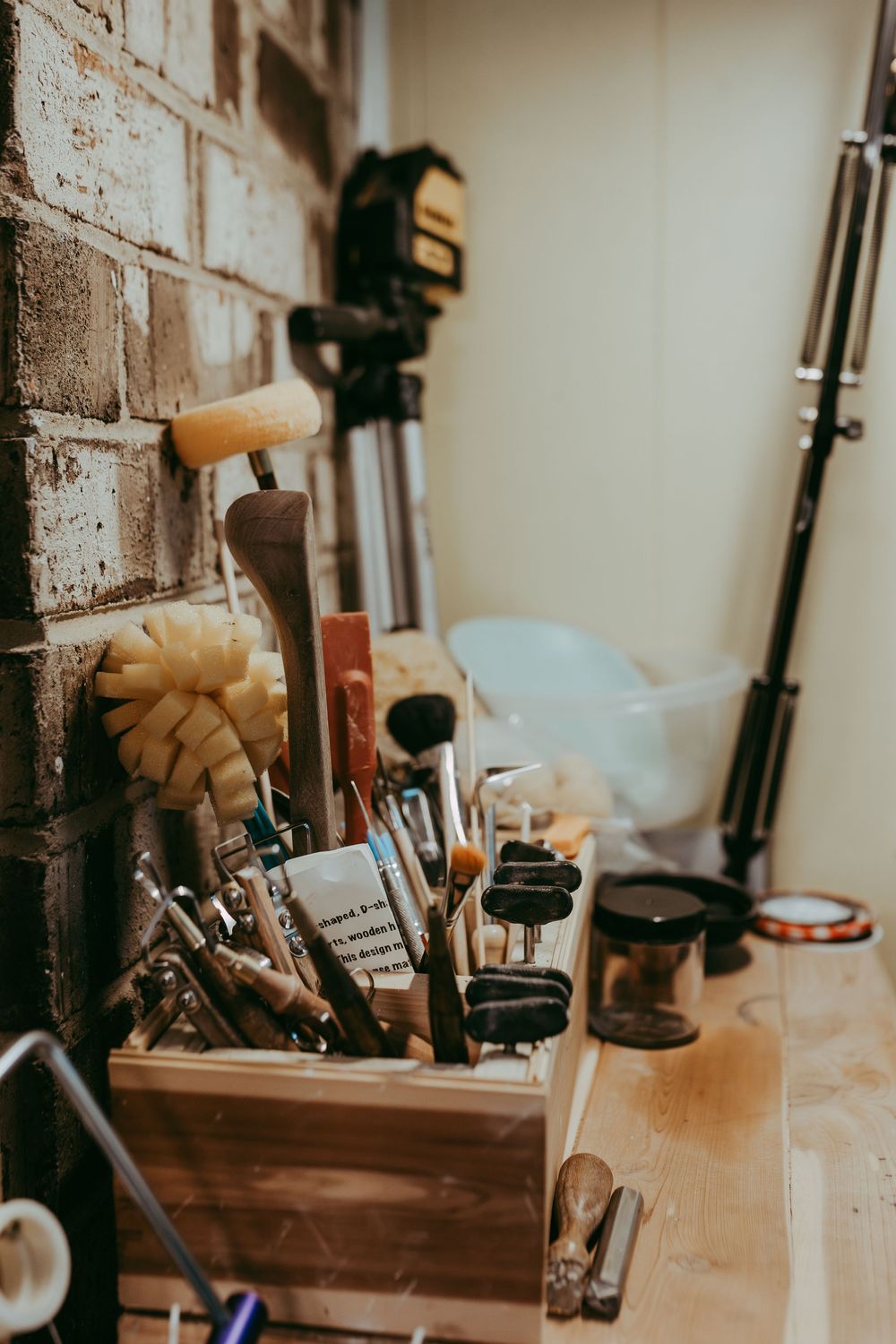 Wooden organizer filled with art tools and paintbrushes on a table near exposed brick wall and photography equipment. Whitney Popp