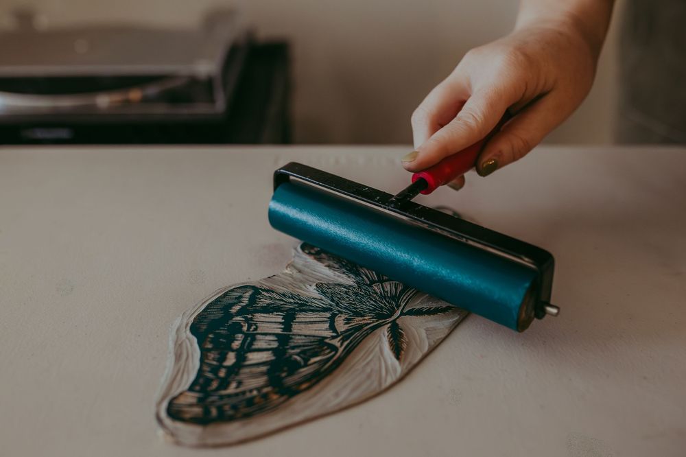 Artist using a teal brayer roller to apply ink onto a butterfly linocut print on a white surface.