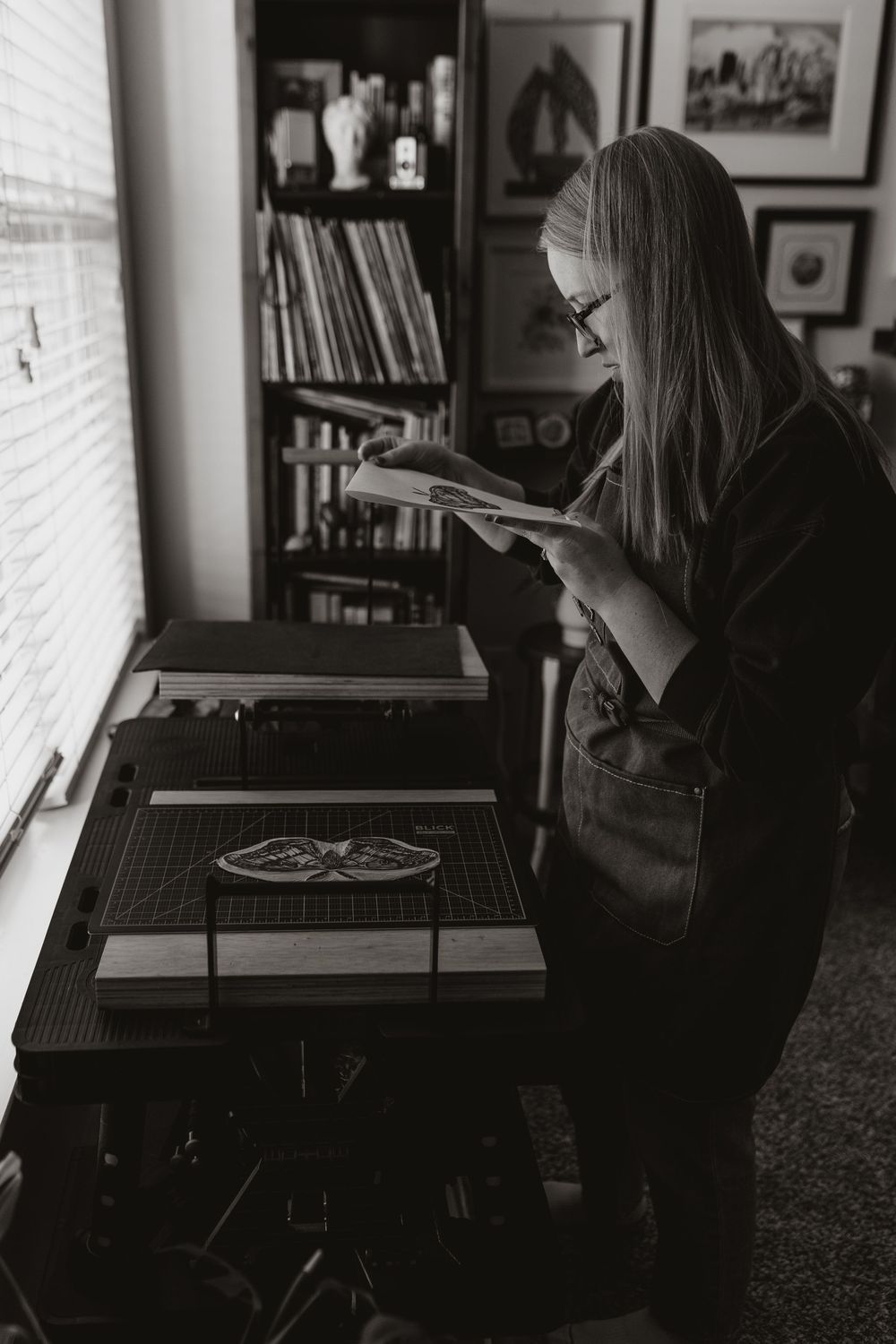 Black and white photo of a woman with long hair reading examining a print beside a worktable in a cozy studio room.