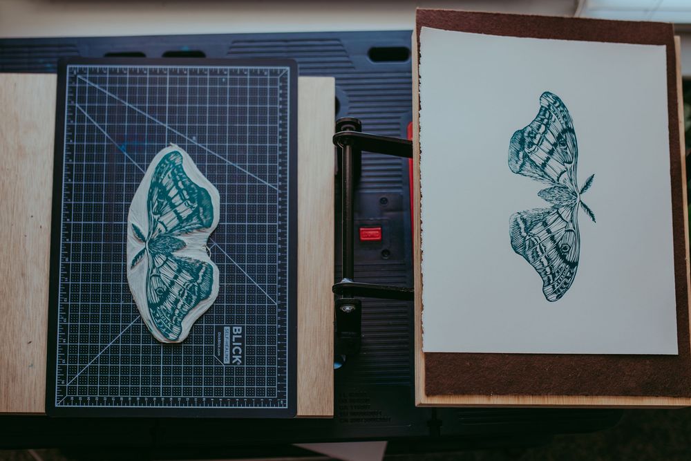 Two moth illustrations on a cutting mat and framed print, featuring teal ink artwork in a printmaking studio setup.