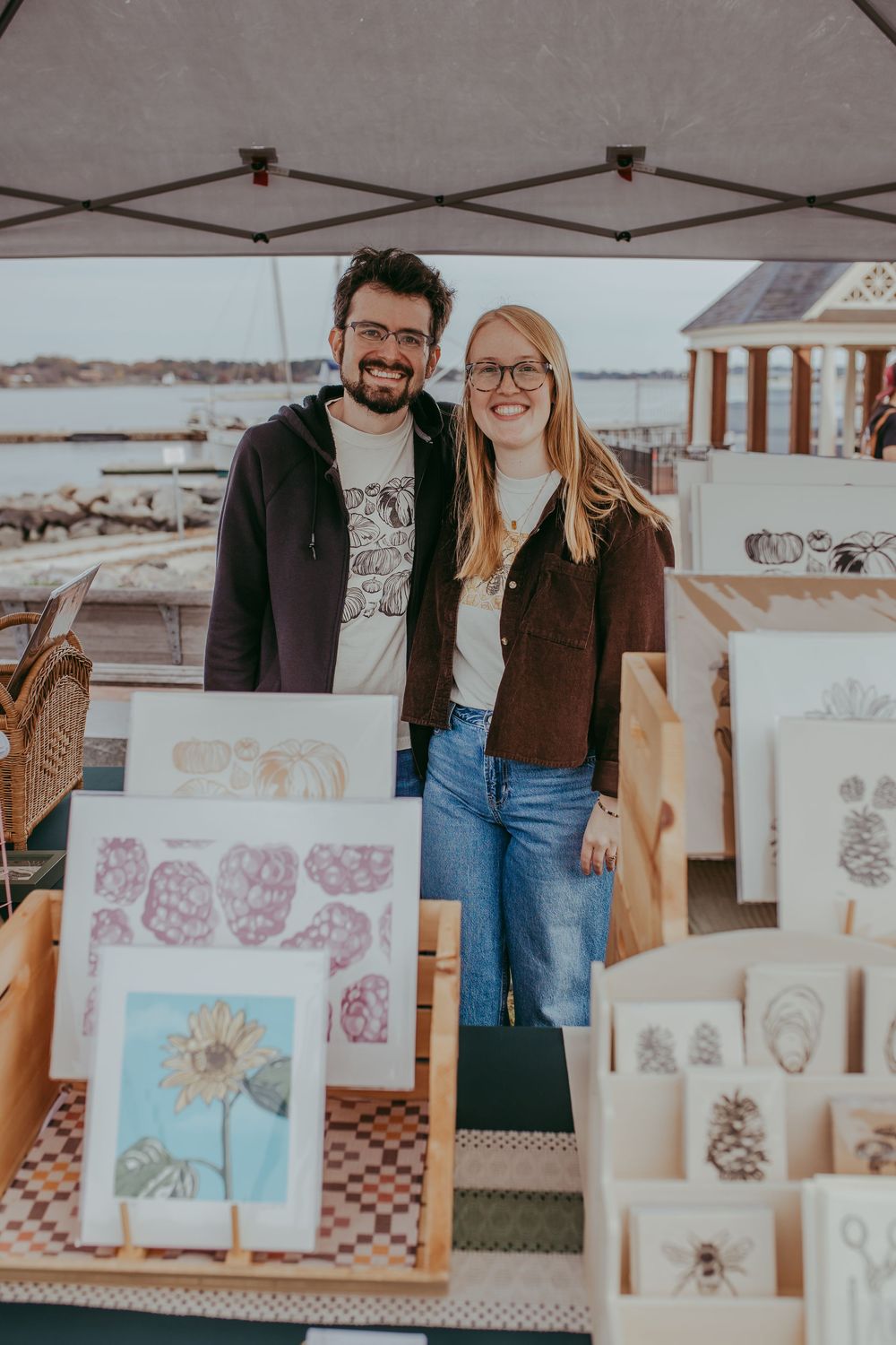 Two smiling artists stand at an outdoor market booth displaying framed botanical prints and nature-themed artwork by the waterfront.