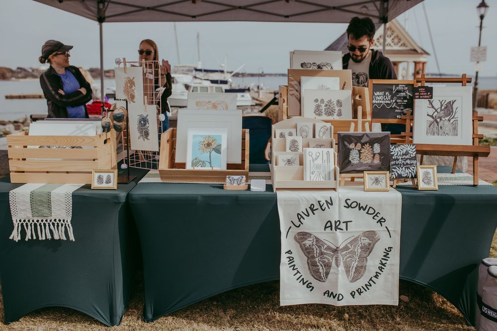 Outdoor art market booth displaying Lauren Bowden Art prints and paintings on tables under a tent near a marina.