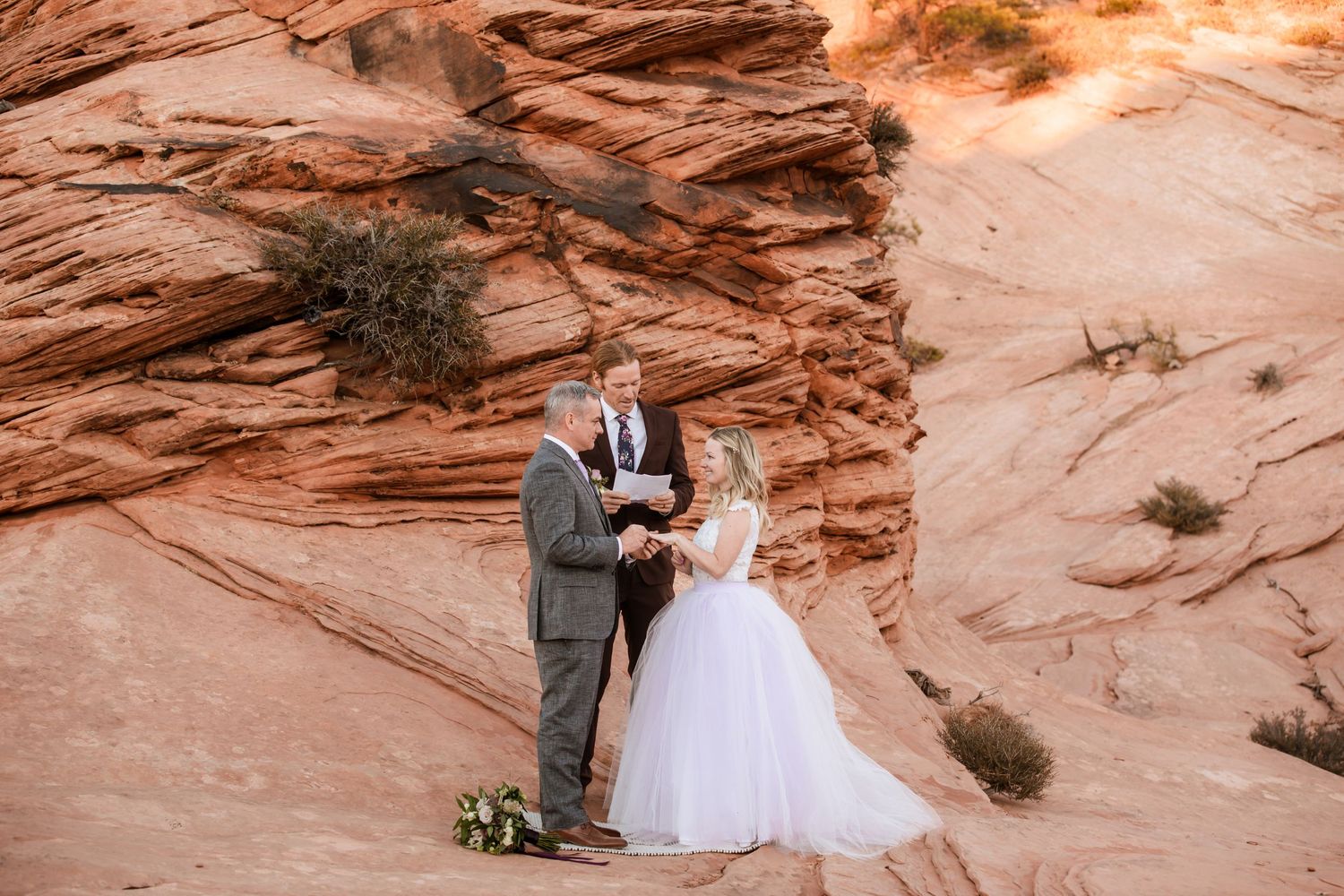 A couple in wedding attire stand together against stunning red rock formations in a desert landscape at sunset.