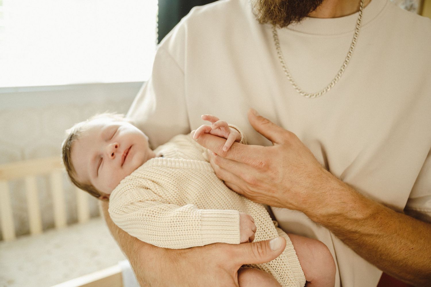 Parent holding sleeping newborn baby wearing knit onesie in natural window light.