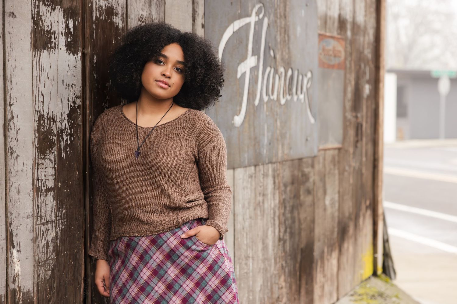 A woman in a brown sweater and plaid skirt poses casually against a rustic wall with graffiti.