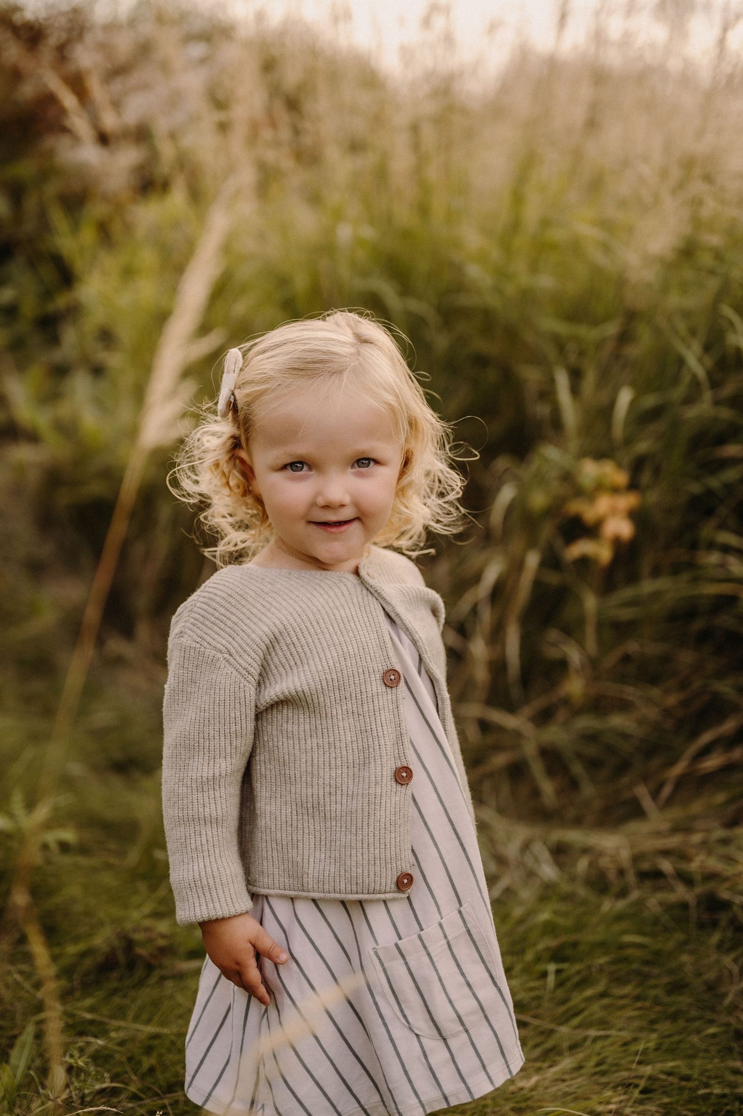 Young child in a striped dress and sweater playing in tall grass.
