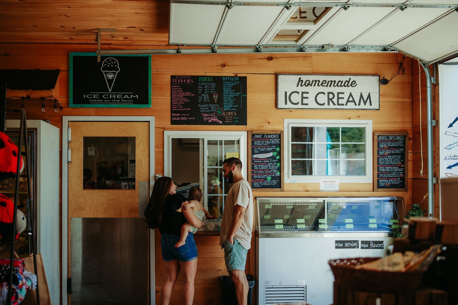 Customers wait at a rustic ice cream shop counter with menu boards and wooden walls.