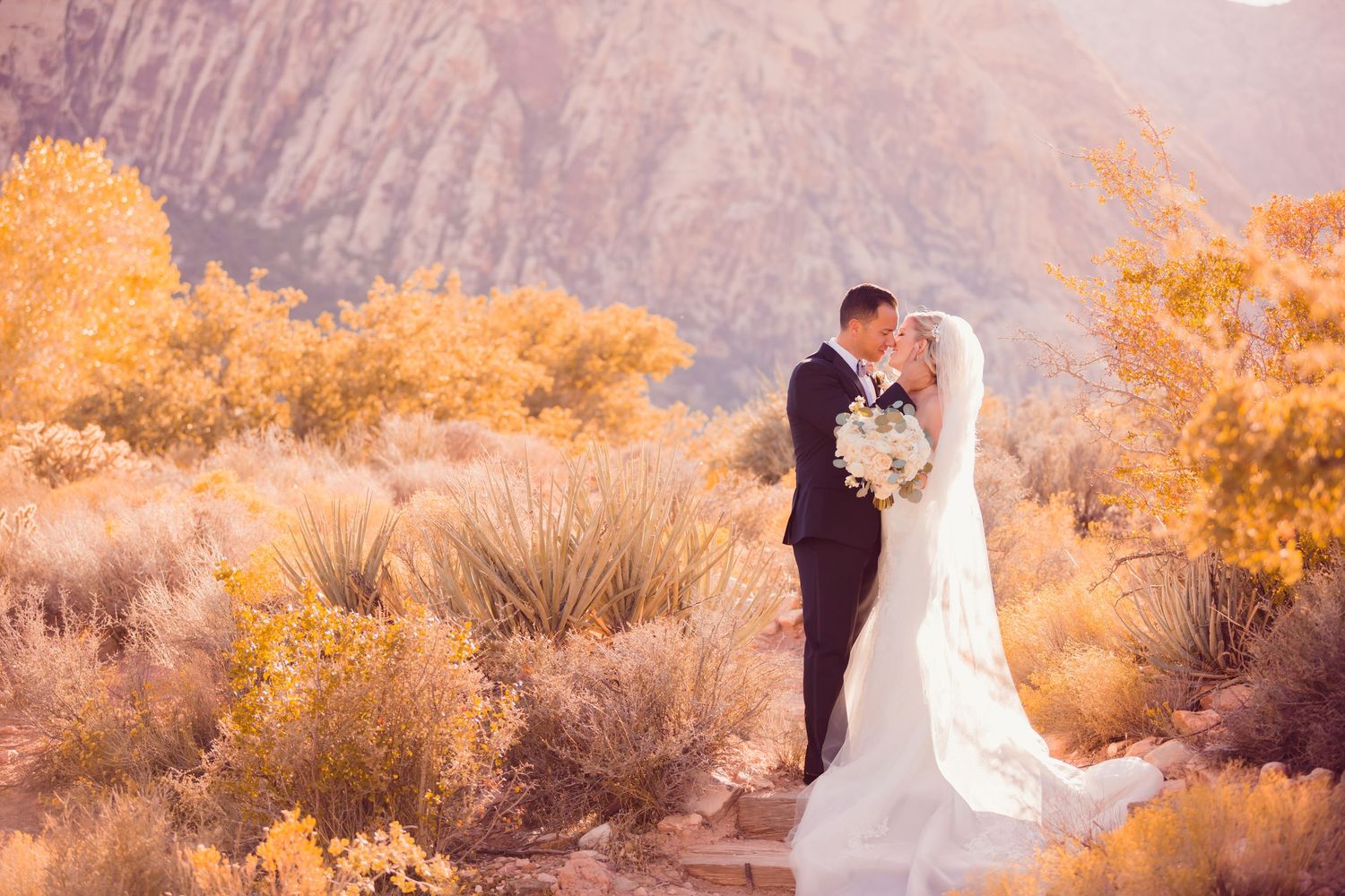 A couple in formal wedding attire walks together through a lush garden path surrounded by trees.
