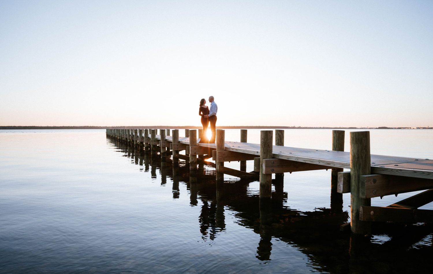 A couple sits on a wooden pier at sunset, silhouetted against the orange sky reflecting on calm waters.
