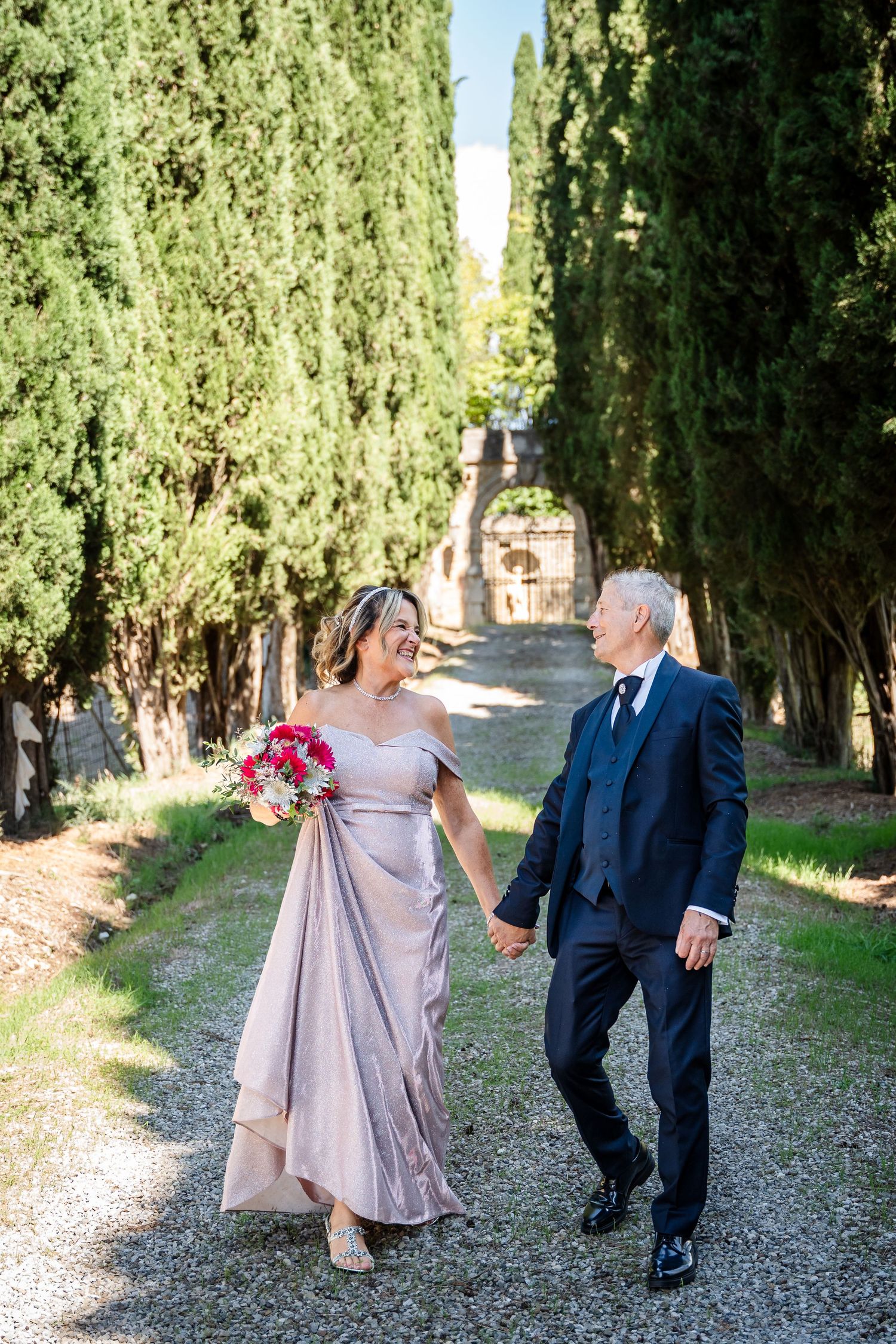 A couple walks hand in hand down a cypress tree-lined gravel path in elegant formal attire.