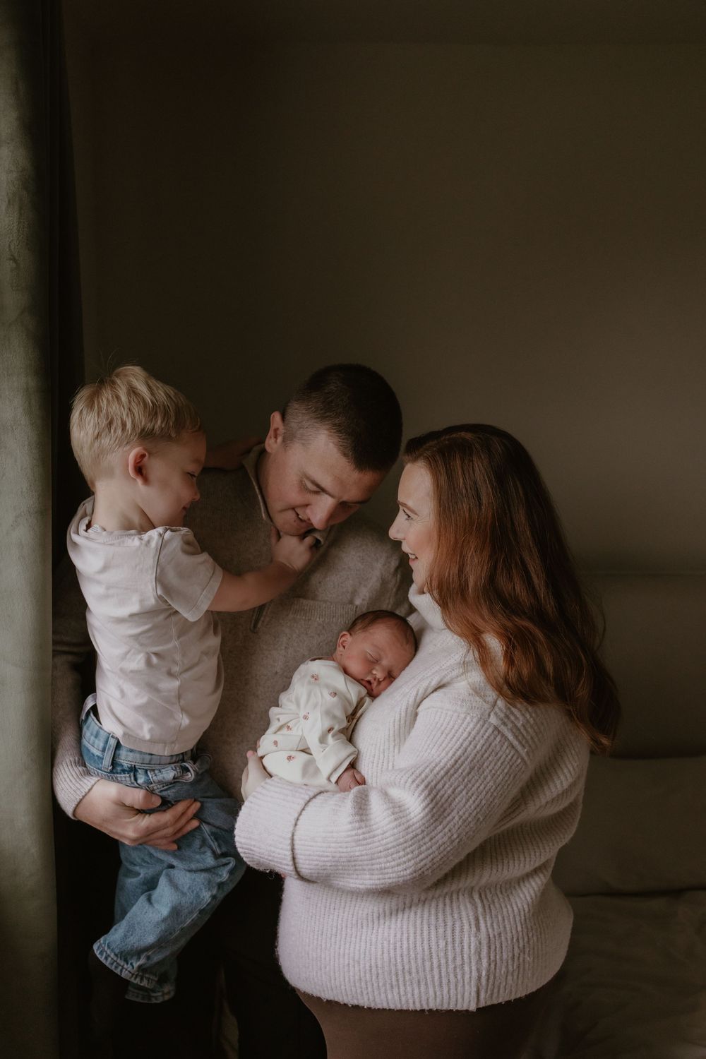 Family shares tender moment with newborn and toddler sibling in muted lighting.