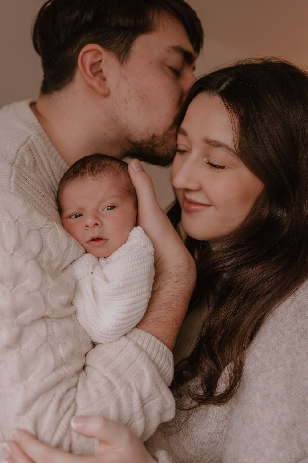 A tender family moment with parents cuddling their newborn baby in white knit clothing.