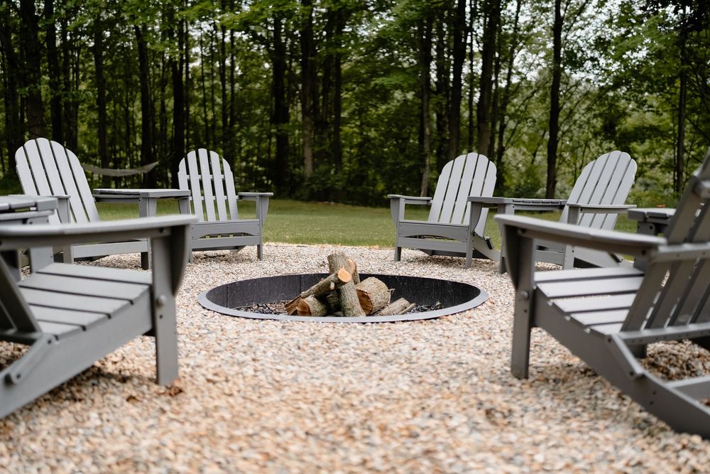 White Adirondack chairs arranged around a modern stone fire pit on a pebbled patio area in a wooded setting.