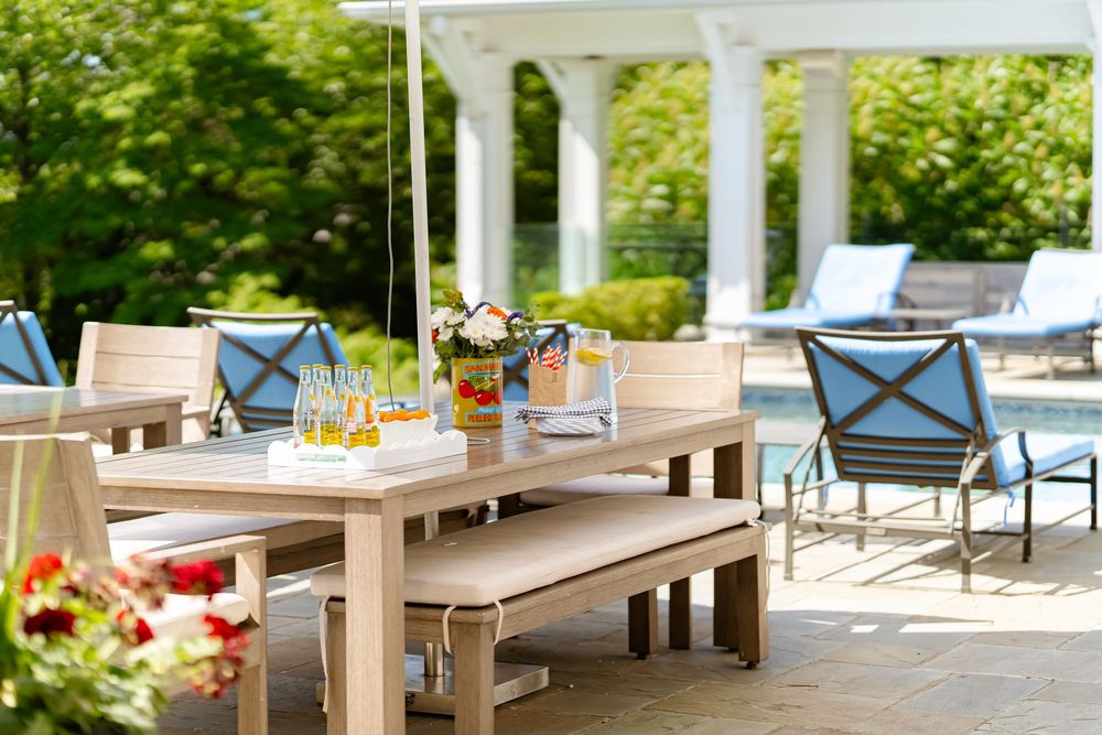 Modern outdoor patio furniture with wooden benches and blue cushioned chairs arranged around tables in a sunny garden setting.