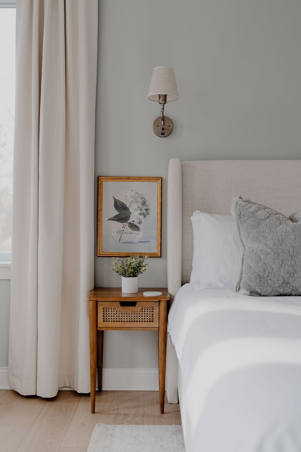 Bright and airy bedroom interior with sage green walls, white curtains, and minimalist botanical artwork in gold frames.