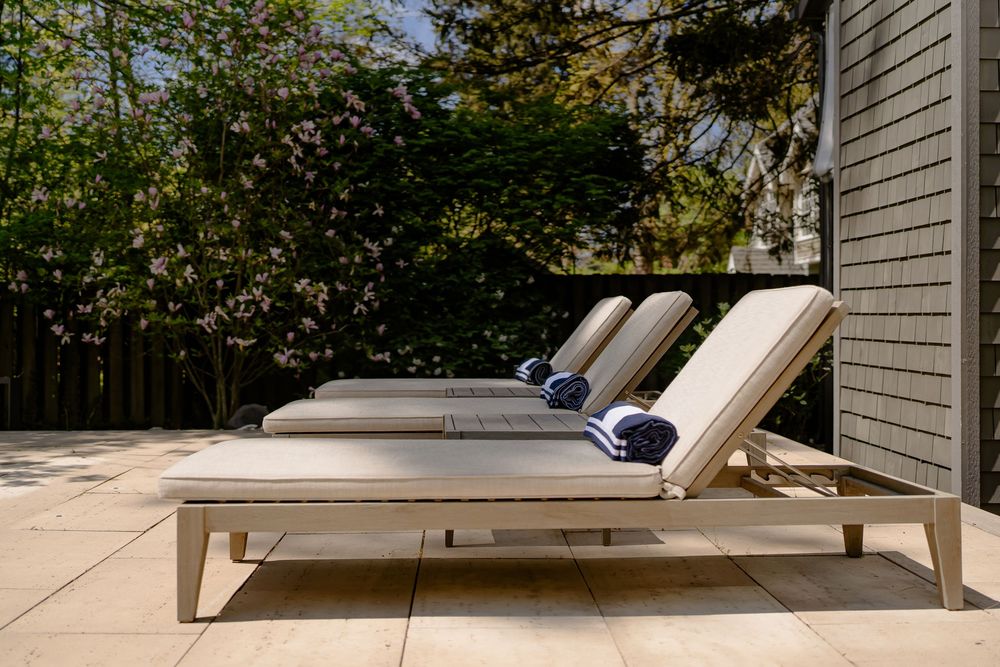 Two modern outdoor chaise lounge chairs with navy cushions on a concrete patio next to a gray house exterior.