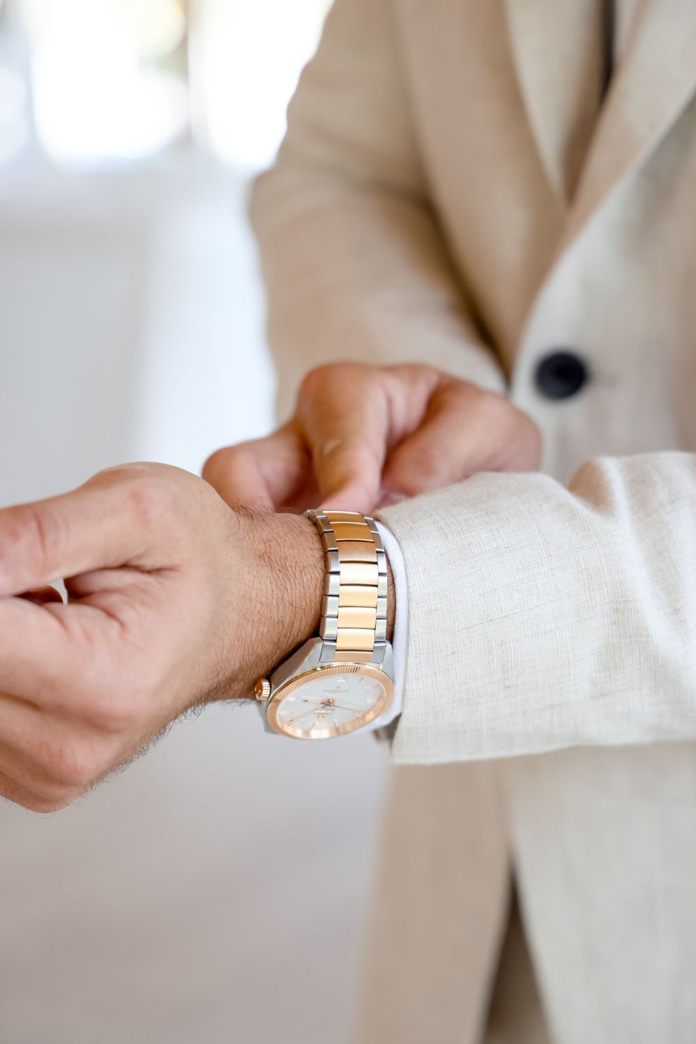 Close-up of hands adjusting a gold and silver two-tone watch with a cream suit sleeve visible.
