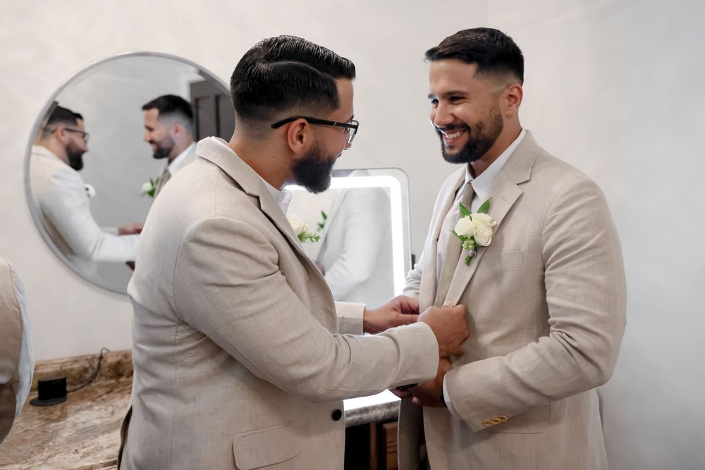 Groomsmen in light colored suits help each other with final wedding day preparations.