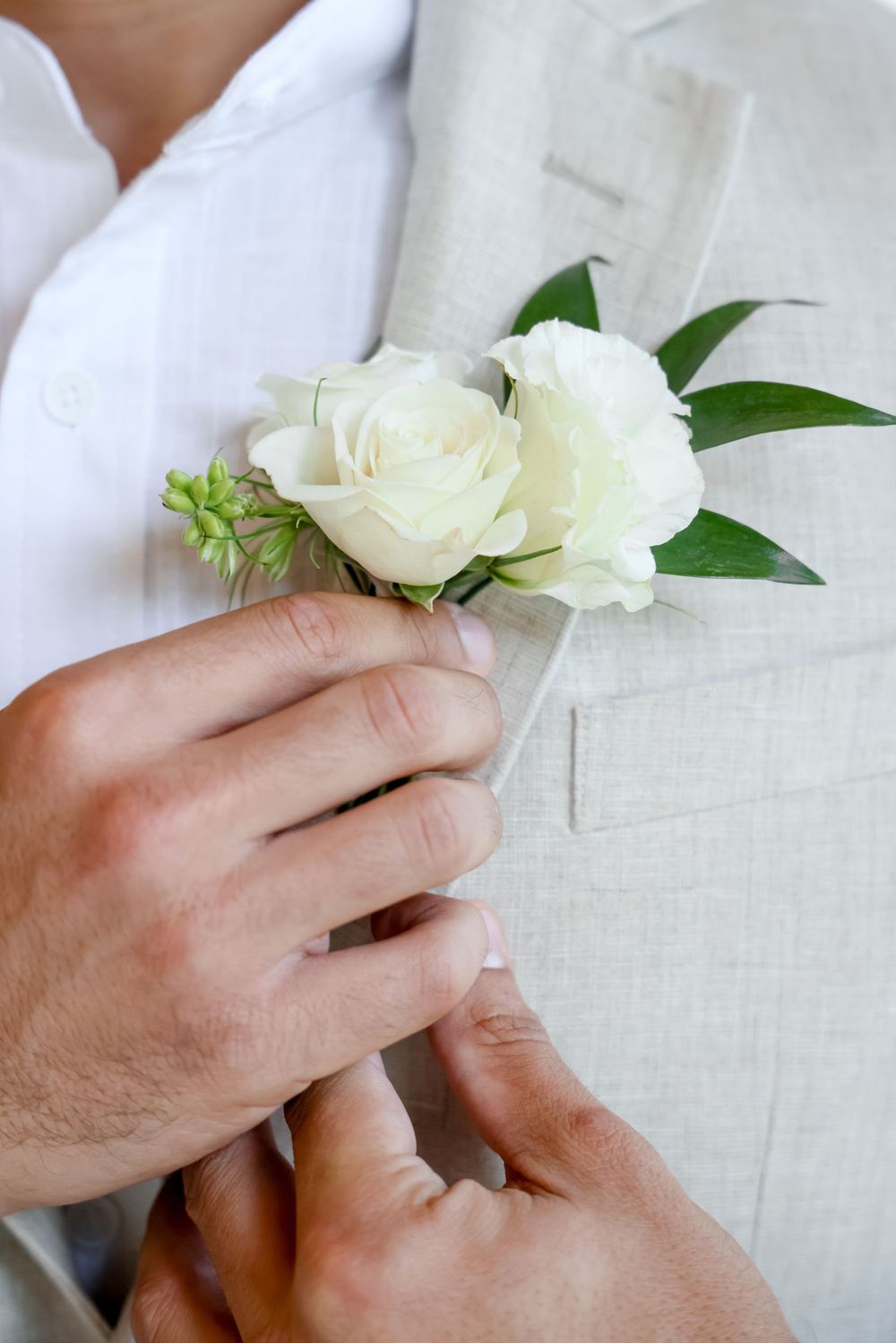 Close-up of hands pinning a white rose boutonniere onto a light colored suit jacket lapel.