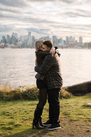Two people embrace warmly on a grassy hill overlooking a city skyline and waterfront at sunset.