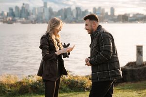 A couple shares a romantic moment along the waterfront with a city skyline visible in the background.