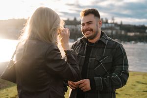 Couple laughing and embracing outdoors against waterfront backdrop at sunset.