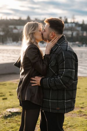A couple shares a romantic kiss at sunset on a grassy waterfront area with a scenic backdrop.