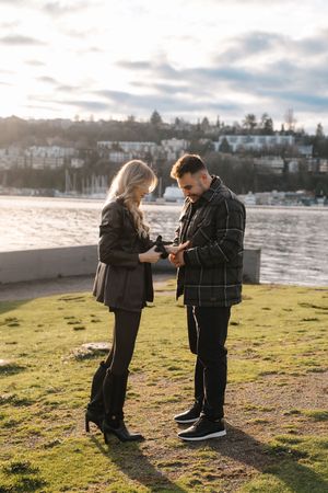 Two people standing by a waterfront on grass looking at their phones during golden hour with houses on a hill in background.