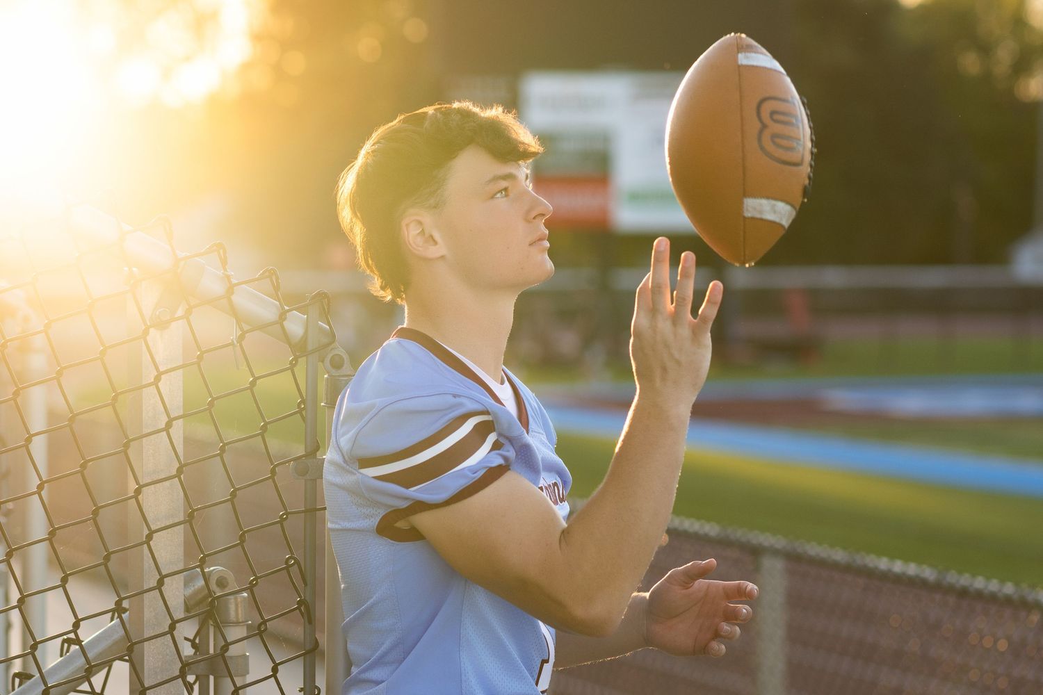Senior story driven photo of a boy tossing his football into the air while the golden sun sets over his football field in the background.