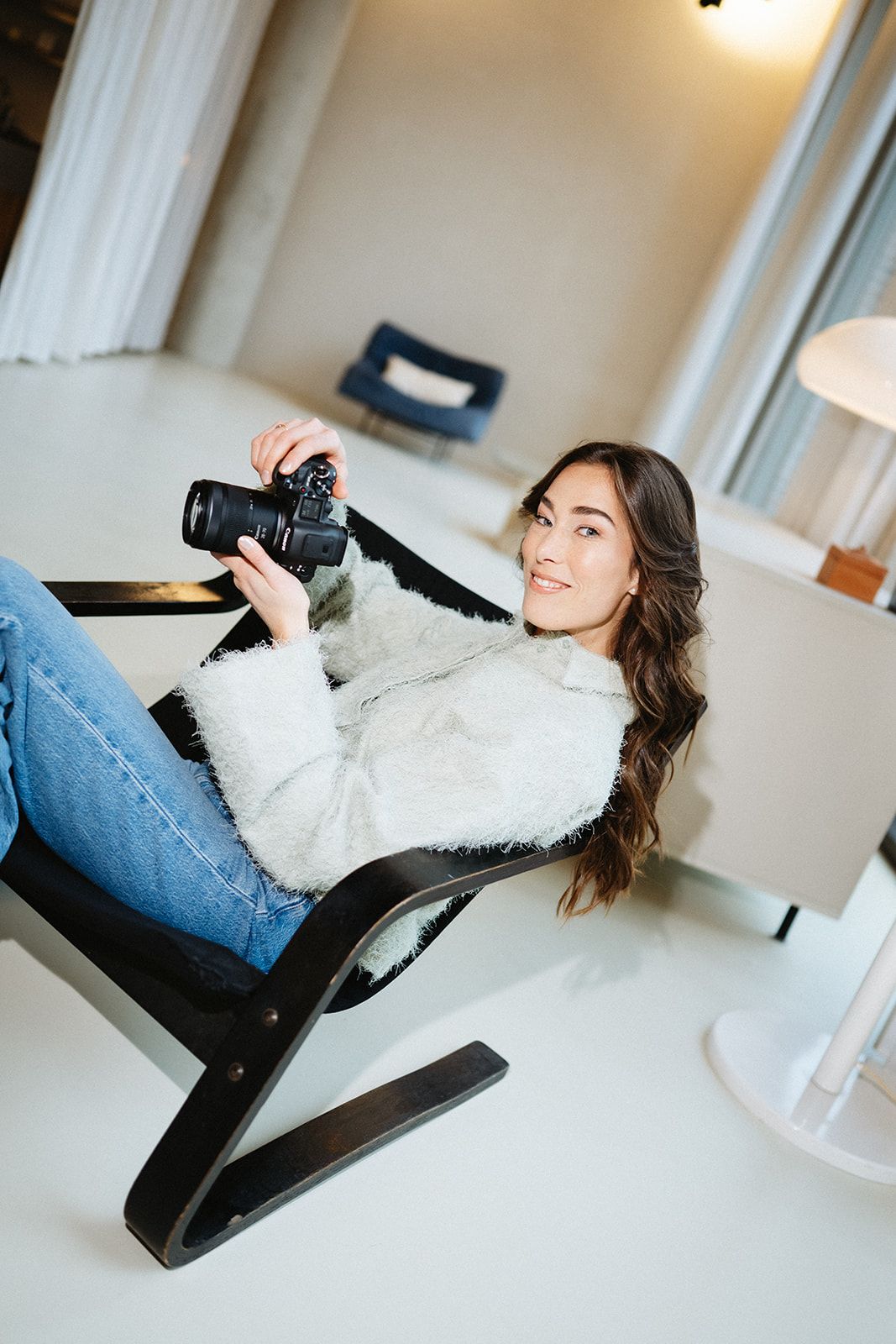 Person in fuzzy white sweater and jeans reclines on chair while holding camera.