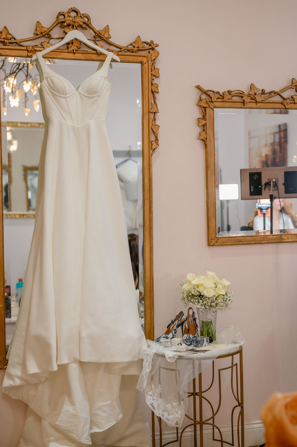 Elegant white wedding dress hanging on display with ornate gold-framed mirrors and floral arrangements.