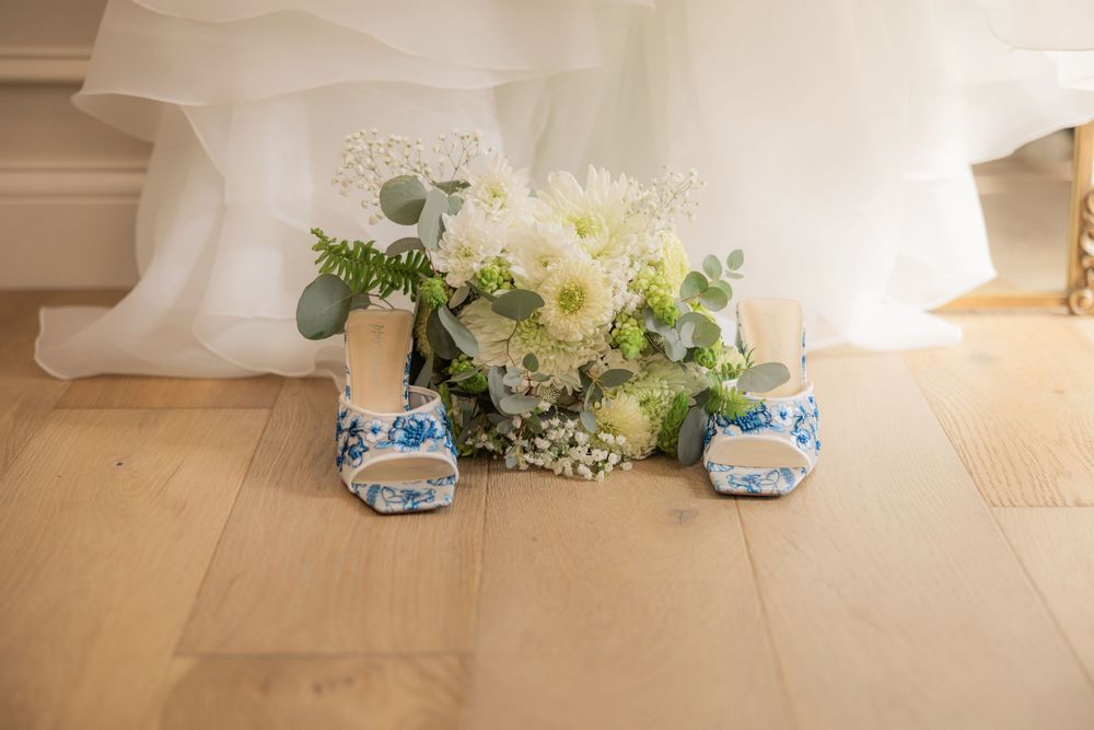 White floral bouquet with greenery sits beside blue and white patterned shoes on a wooden floor.