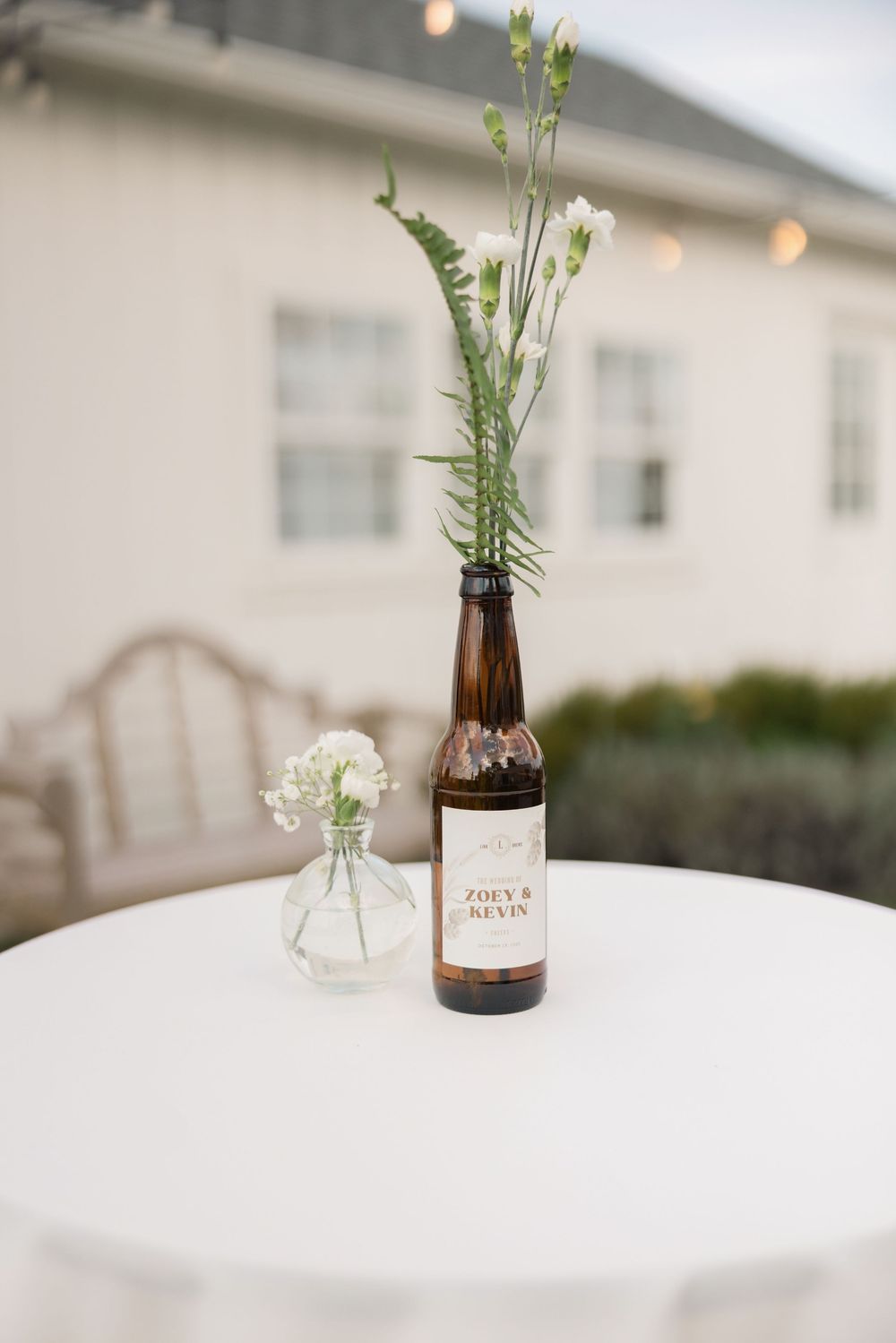 A brown beer bottle with delicate white flowers serves as a rustic vase centerpiece on a white round table. aspen and alston ohio