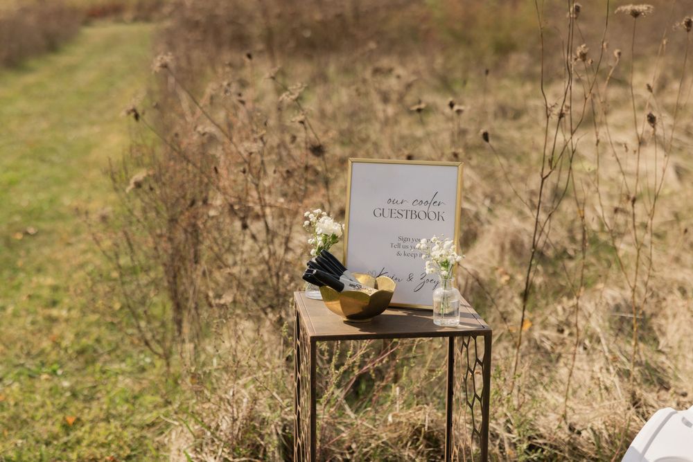 A rustic metal table displays a white ceremony sign surrounded by dried wildflowers in a golden field.