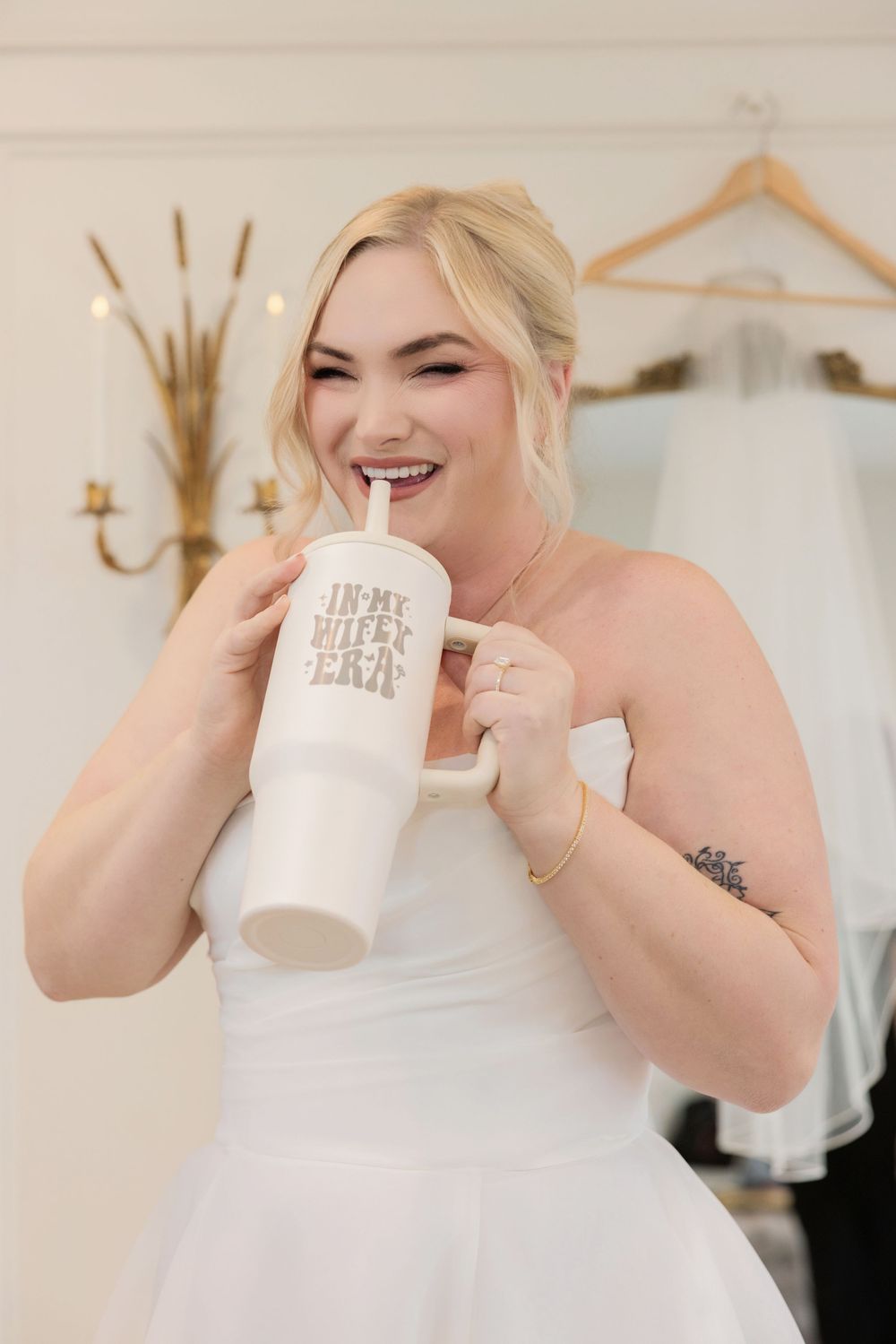 Someone in a white wedding dress sips from a large white tumbler cup while smiling.