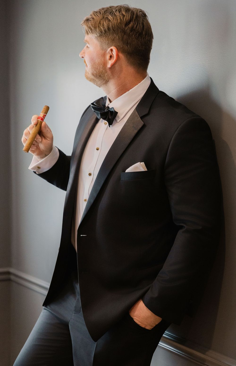 A person in formal black tuxedo and bow tie holds a snack while posing against a gray wall.