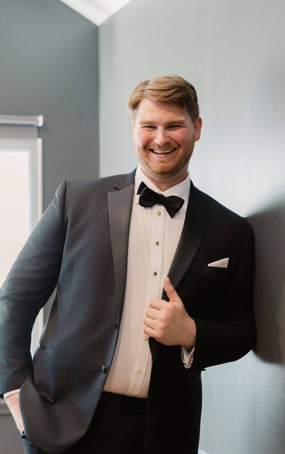 A groom in a classic black tuxedo with bow tie smiles while adjusting his jacket in a room with blue-gray walls.
